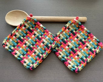 Pode incluir: Duas pegas coloridas tecidas com uma colher de pau sobre uma superfície cinza. As pegas são quadradas e apresentam uma mistura vibrante de fios rosa, laranja, verde, azul e branco. Cada pega tem uma argola para pendurar.