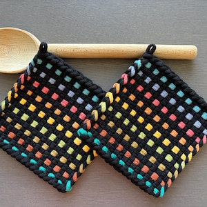 Könnte beinhalten: Zwei schwarze quadratische Topflappen mit einem gewebten Design mit bunten Quadraten in Rot, Orange, Gelb, Grün und Blau. Ein Holzlöffel liegt über den Topflappen. Die Topflappen haben eine Schlaufe zum Aufhängen.