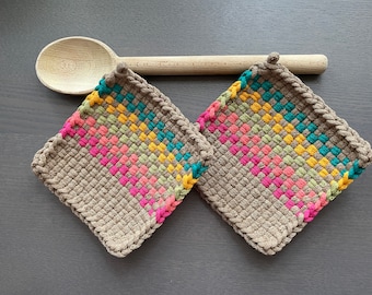 Pode incluir: Duas pegas quadradas tecidas com um padrão geométrico colorido em rosa, amarelo, verde e turquesa. As pegas são de cor bege neutra e têm uma borda trançada. Uma colher de pau repousa ao fundo.