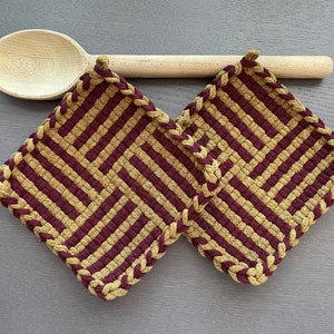 May include: Two square, woven pot holders with a geometric pattern in burgundy and tan. The edges are braided in tan. A wooden spoon rests above the pot holders on a grey surface. Kitchen and dining decor.