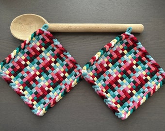 Pode incluir: Duas pegas tecidas em padrão de losango, com uma mistura de fios vermelhos, verde-água, rosa, amarelo e branco. Cada pega tem uma argola para pendurar. Uma colher de pau repousa por cima.