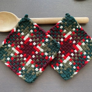 May include: Two square woven potholders with a plaid pattern in red, white, green, and burgundy. The potholders are made of a thick, textured material and have a loop for hanging. A wooden spoon rests behind the potholders.
