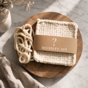May include: A stack of off-white, square, textured crochet coasters wrapped in a brown paper band with a white question mark and the text mystery set printed on it, accompanied by a loose, coiled off-white crochet cord.