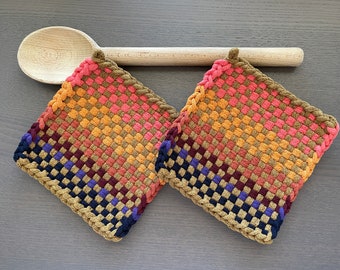 May include: Two square woven potholders in a gradient of colors, including pink, orange, brown, and blue. The potholders have a braided border and a loop for hanging. A wooden spoon rests above the potholders.