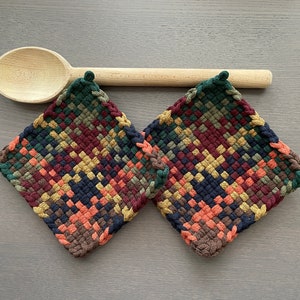 May include: Two square woven potholders in a diamond pattern with a mix of colors including green, burgundy, orange, and yellow. A wooden spoon rests above the potholders. The potholders are approximately 7 inches square.