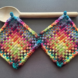 Puede incluir: Dos salvamanteles cuadrados tejidos con un arcoíris de colores, incluyendo azul, verde, amarillo, naranja y morado. Una cuchara de madera descansa encima. Cada salvamanteles tiene un lazo para colgar.