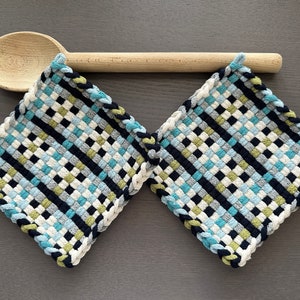May include: Two woven pot holders in a blue, green, black, and white checkered pattern with a rope-like border. The pot holders are hanging on a wooden spoon.