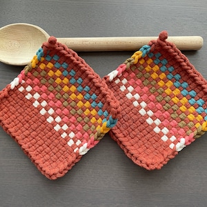 Op de afbeelding: Twee vierkante, geweven pannenlappen in een roestkleur, met een kleurrijk geometrisch patroon van wit, roze, geel, blauw en bruin. Een houten lepel rust boven de pannenlappen. Keukenaccessoires.
