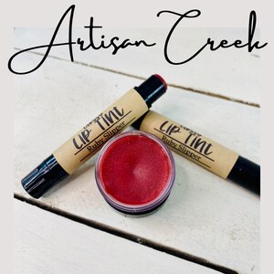 May include: Two lip tints in the shade "Ruby Slipper" by Artisan Creek. One is a tube with a black cap and the other is a small jar with a red lip tint inside.
