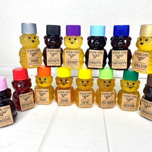 May include: A collection of 15 small glass bear-shaped bottles filled with honey. The bottles are arranged in three rows on a white wooden surface. Each bottle has a different flavor of honey, including lavender, blueberry, vanilla bean, peach, lemon, garlic, apple caramel, jalapeno, and chocolate.
