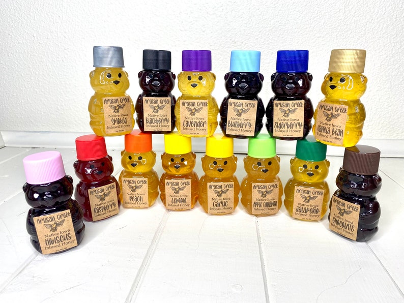 May include: A collection of 15 small glass bear-shaped bottles filled with honey. The bottles are arranged in three rows on a white wooden surface. Each bottle has a different flavor of honey, including lavender, blueberry, vanilla bean, peach, lemon, garlic, apple caramel, jalapeno, and chocolate.