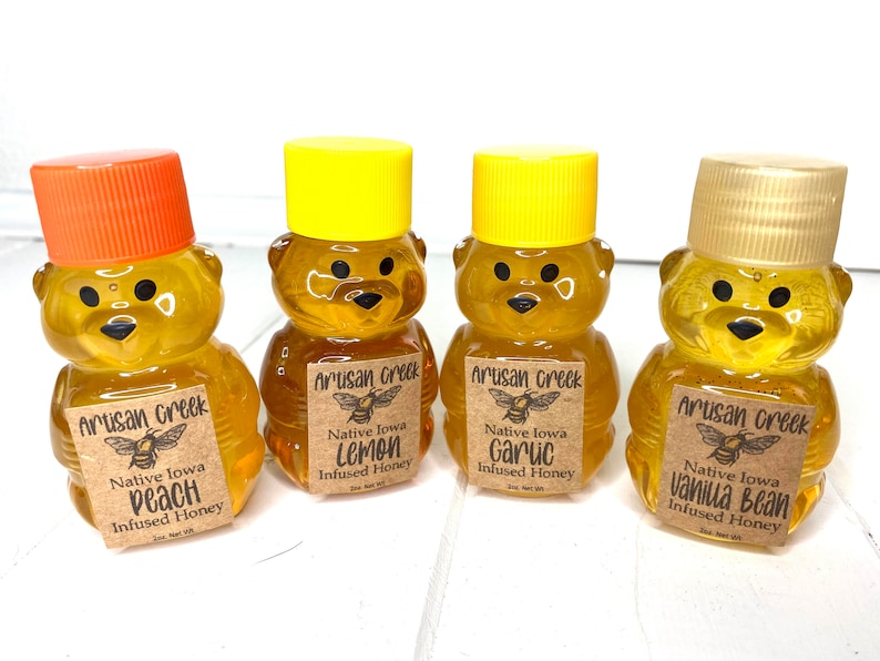May include: Four small honey bear-shaped bottles filled with honey. Each bottle has a brown paper label with black text that reads "Artisan Creek", "Native Iowa", and the flavor of the honey. The flavors are Peach, Lemon, Garlic, and Vanilla Bean.