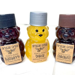 May include: Three small glass bear-shaped bottles filled with honey. The bottles are labeled with brown paper labels that say "Artisan Creek" and list the honey flavors: "Blackberry", "Smoked", and "Chocolate".