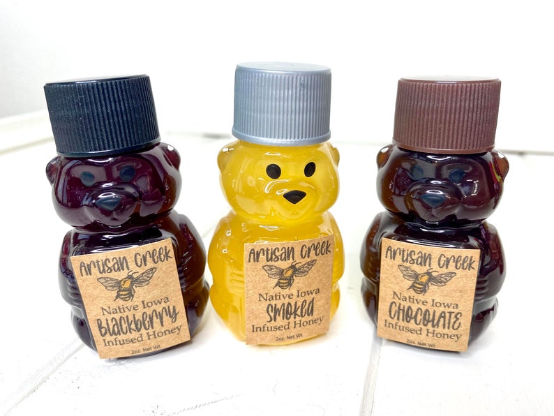 May include: Three small glass bear-shaped bottles filled with honey. The bottles are labeled with brown paper labels that say "Artisan Creek" and list the honey flavors: "Blackberry", "Smoked", and "Chocolate".