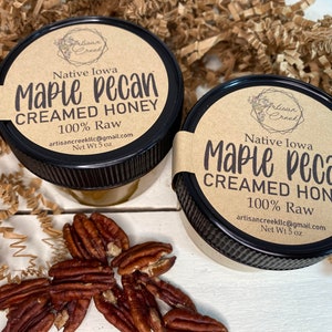 May include: Two small glass jars with black lids filled with creamed honey. The labels are brown with black text and say "Native Iowa Maple Pecan Creamed Honey 100% Raw artisancreeklic@gmail.com Net Wt 5 oz". There are pecan halves scattered on a white surface in front of the jars.