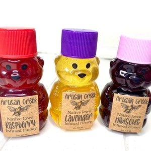 May include: Three small bear-shaped bottles of honey, each with a different flavor: raspberry, lavender, and hibiscus. The bottles are labeled with the text "Artisan Creek" and "Native Iowa" along with the flavor.