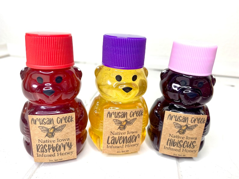 May include: Three small bear-shaped bottles of honey, each with a different flavor: raspberry, lavender, and hibiscus. The bottles are labeled with the text "Artisan Creek" and "Native Iowa" along with the flavor.