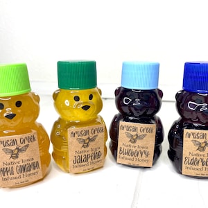 May include: Four small bear-shaped bottles of honey, each with a different flavor: apple cinnamon, jalape&ntilde;o, blueberry, and elderberry. Each bottle has a brown label with the text "Artisan Creek" and the flavor of the honey.