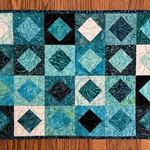 May include: A teal and black quilted table runner with a diamond pattern. The runner is made of fabric with a paisley design.