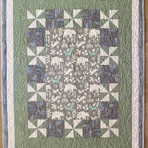 May include: A green and blue baby quilt with a white fabric center featuring a woodland animal print. The quilt is made with a patchwork design and has a white border.