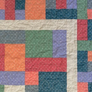 Op de afbeelding: Een patchworkdeken met vierkanten in verschillende kleuren, waaronder rood, oranje, groen, blauw en wit. De vierkanten zijn in een willekeurig patroon aan elkaar gestikt, wat een uniek en kleurrijk ontwerp cre&euml;ert.