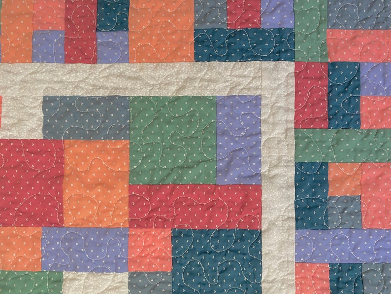 Op de afbeelding: Een patchworkdeken met vierkanten in verschillende kleuren, waaronder rood, oranje, groen, blauw en wit. De vierkanten zijn in een willekeurig patroon aan elkaar gestikt, wat een uniek en kleurrijk ontwerp cre&euml;ert.