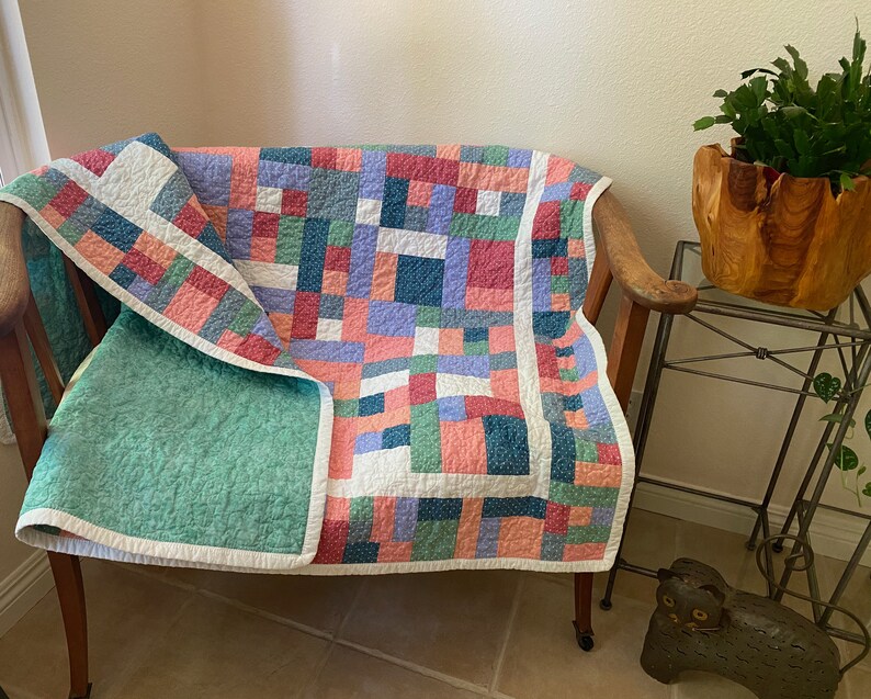 Op de afbeelding: Een patchworkdeken met een groene, roze, blauwe en witte kleurstelling. De deken is gevouwen over een houten stoel met een witte rand.