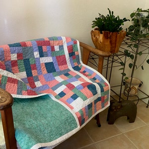 Op de afbeelding: Een patchworkdeken in tinten roze, blauw, groen en wit is over een houten fauteuil gelegd. De deken heeft een eenvoudig, geometrisch patroon en is gemaakt van verschillende stoffen. De fauteuil staat in een woonkamer met een plantenstandaard en een potplant.