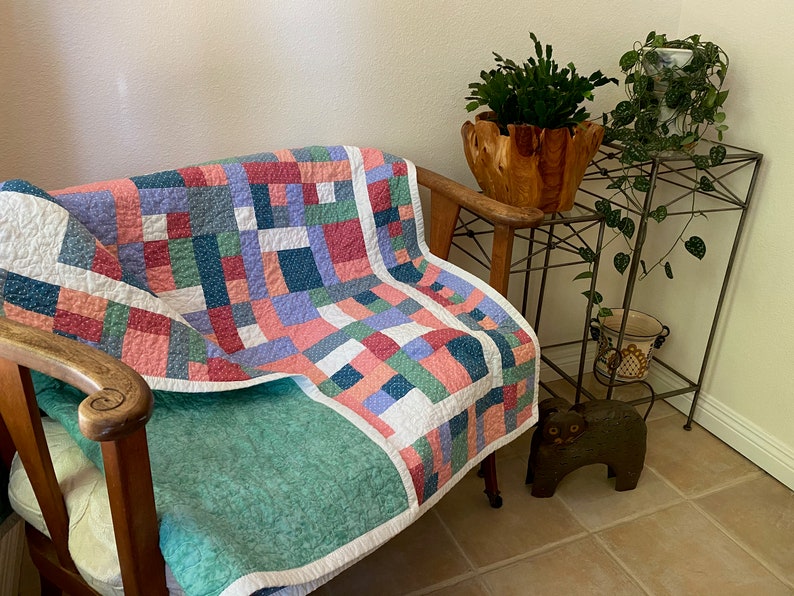 Op de afbeelding: Een patchworkdeken in tinten roze, blauw, groen en wit is over een houten fauteuil gelegd. De deken heeft een eenvoudig, geometrisch patroon en is gemaakt van verschillende stoffen. De fauteuil staat in een woonkamer met een plantenstandaard en een potplant.