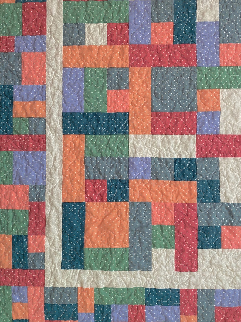 Op de afbeelding: Een patchworkdeken met een verscheidenheid aan kleuren en patronen, waaronder rood, oranje, groen, blauw en paars. De deken is gemaakt van vierkanten en rechthoeken, en de naden zijn zichtbaar. De deken is gestikt met een eenvoudig steekpatroon.