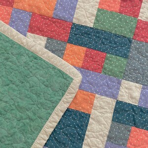 Op de afbeelding: Een patchworkdeken met verschillende kleuren en patronen, waaronder groen, blauw, paars en oranje. De deken is gemaakt van vierkanten en rechthoeken, en de naden zijn zichtbaar. De deken is dubbelgevouwen, en de rand van de deken is zichtbaar.