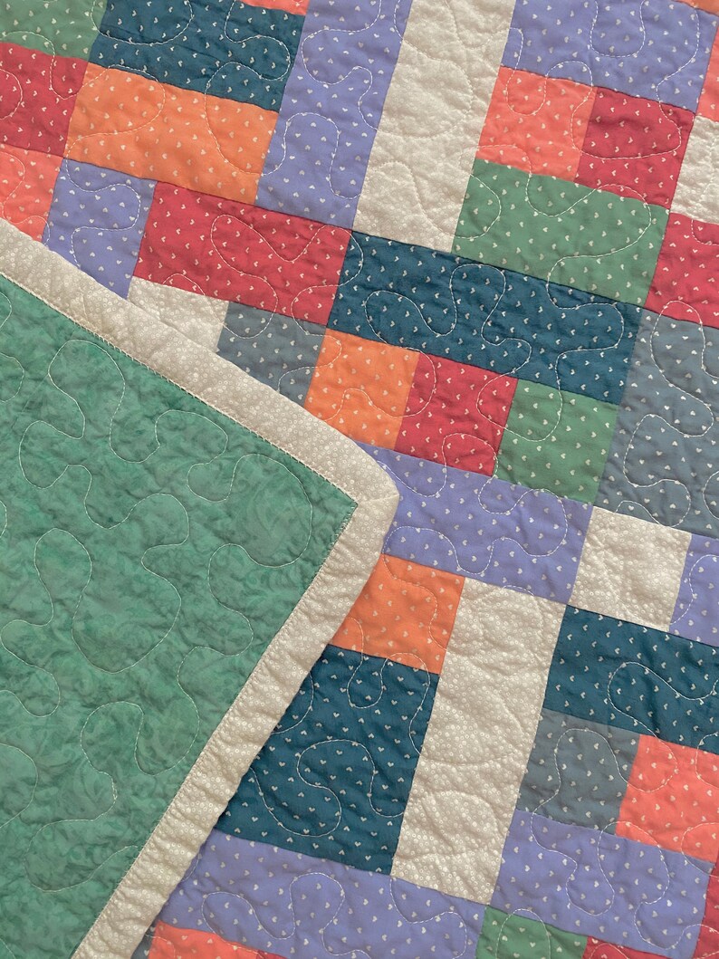 Op de afbeelding: Een patchworkdeken met verschillende kleuren en patronen, waaronder groen, blauw, paars en oranje. De deken is gemaakt van vierkanten en rechthoeken, en de naden zijn zichtbaar. De deken is dubbelgevouwen, en de rand van de deken is zichtbaar.