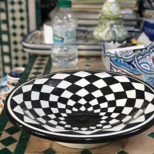Peut inclure: Un bol en céramique noir et blanc avec un motif à carreaux. Le bol a un bord large et un intérieur profond et arrondi. Le motif crée un effet d'illusion d'optique tourbillonnant.