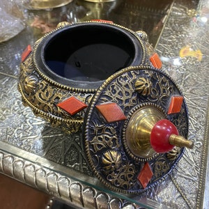 May include: A decorative box with a lid, featuring intricate gold and black designs and red accents. The box is made of metal and has a round shape.