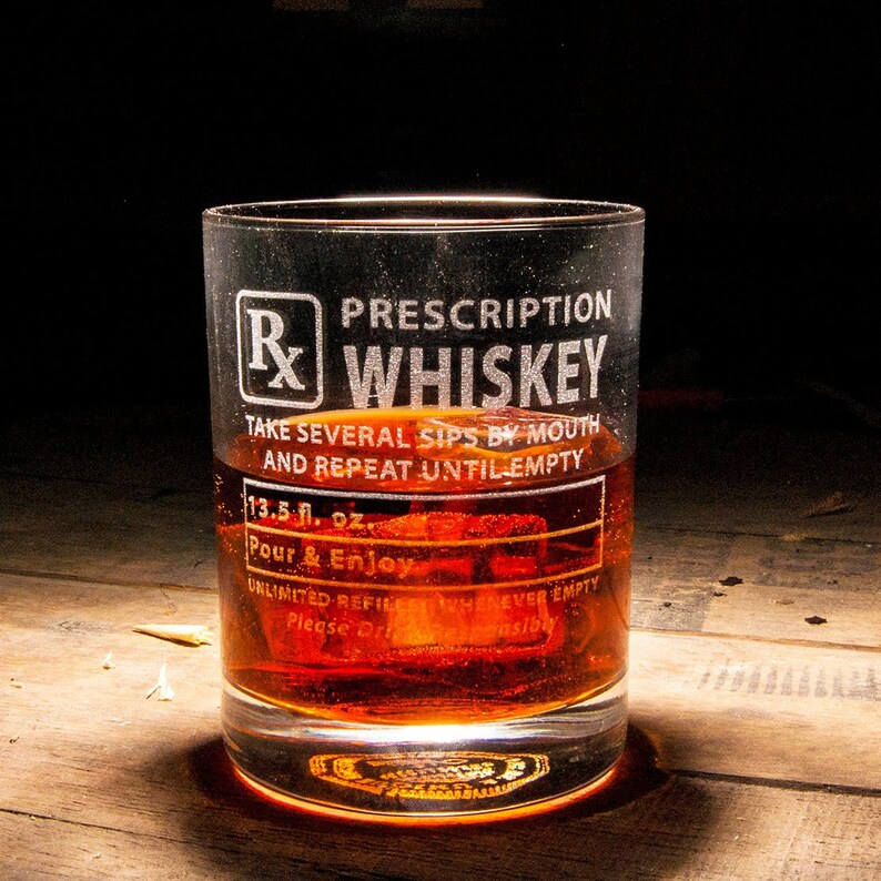 Op de afbeelding: Een heldere glazen whiskey tumbler met een wit ge&euml;tst ontwerp met de tekst "Prescription Whiskey" en "Neem meerdere slokjes door de mond en herhaal tot het leeg is". Het glas is gevuld met amberkleurige whiskey en ijs.