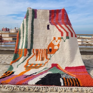 May include: A white, hand-woven rug with a geometric pattern in shades of orange, pink, green, and black. The rug has a fringe border and is displayed on a rooftop.