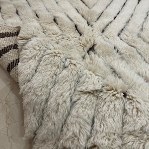 K&ouml;nnte beinhalten: Ein wei&szlig;er und brauner Shaggy-Teppich mit geometrischem Muster. Der Teppich hat einen dicken Flor und ist aus Naturfasern gefertigt.