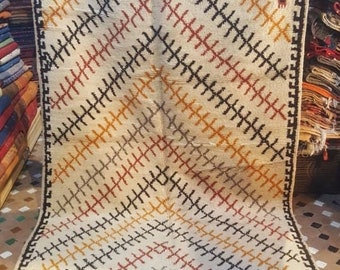 May include: A white wool rug with a geometric pattern in black, red, orange, and gray. The rug has a fringe border.