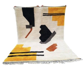 Könnte beinhalten: Ein weißer Teppich mit einem geometrischen Muster in Schwarz, Grau, Braun und Gelb. Der Teppich hat einen Fransenrand.