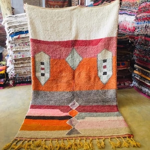 May include: A colorful Moroccan rug with a geometric pattern in shades of orange, red, pink, and gray. The rug has a fringe border and is made of wool.