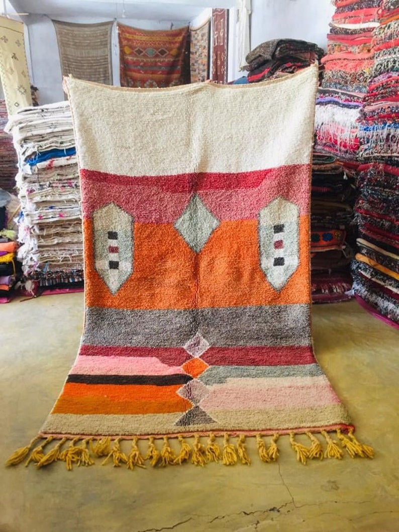 May include: A colorful Moroccan rug with a geometric pattern in shades of orange, red, pink, and gray. The rug has a fringe border and is made of wool.