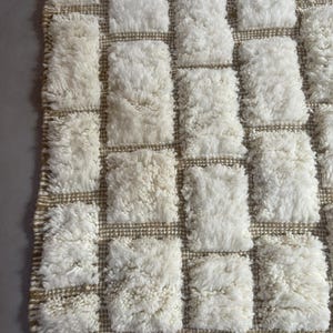 May include: A white and beige woven rug with a checkered pattern. The rug is made of fluffy, textured yarn.