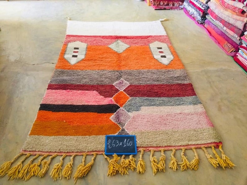 May include: A colorful Moroccan rug with a geometric pattern in shades of orange, pink, red, and gray. The rug has a fringe border and a handwritten sign that reads "2.63 x 1.60".