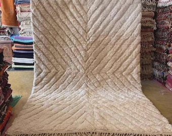 May include: A large, beige rug with a geometric chevron pattern. The rug has a thick, textured pile and a fringe along the bottom edge. The rug is displayed in a shop with other rugs.