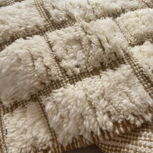 May include: A white and beige woven rug with a textured, fluffy surface. The rug has a geometric pattern with squares.