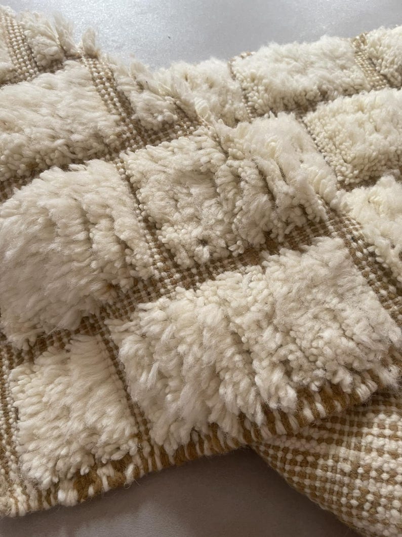 May include: A white and beige woven rug with a textured, fluffy surface. The rug has a geometric pattern with squares.
