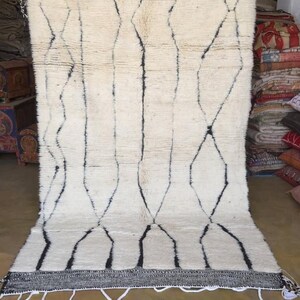 May include: A white wool rug with a black geometric pattern. The rug has a fringe border and is made in Morocco.
