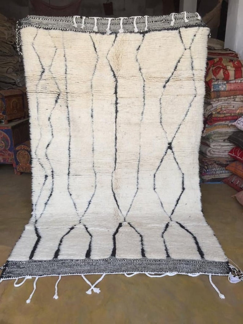May include: A white wool rug with a black geometric pattern. The rug has a fringe border and is made in Morocco.