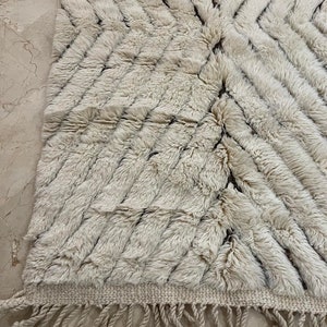 K&ouml;nnte beinhalten: Ein wei&szlig;er und brauner marokkanischer Teppich mit geometrischem Muster und Fransen. Der Teppich ist aus Wolle und hat eine weiche, flauschige Textur.