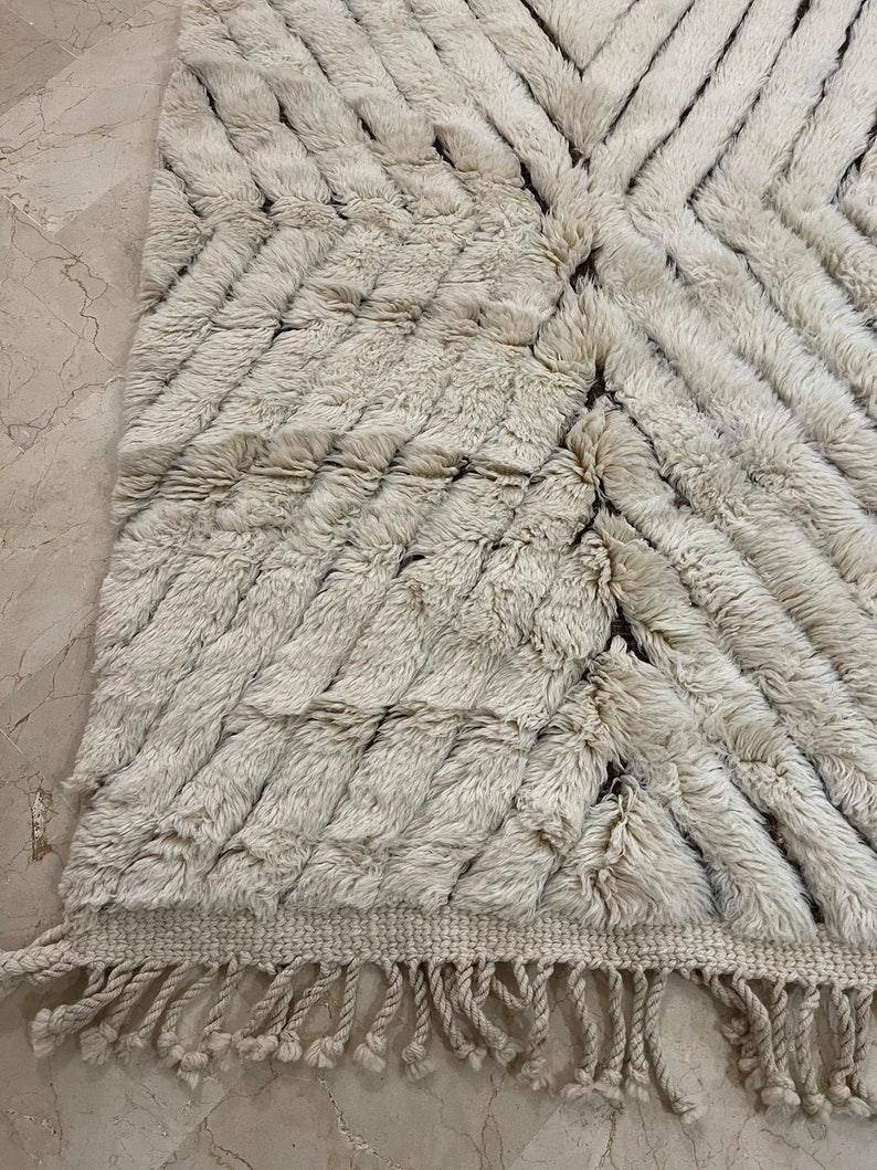 K&ouml;nnte beinhalten: Ein wei&szlig;er und brauner marokkanischer Teppich mit geometrischem Muster und Fransen. Der Teppich ist aus Wolle und hat eine weiche, flauschige Textur.